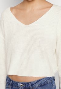 Pull court blanc en maille avec un col en v et une texture côtelée. Associé à un jean en denim bleu avec un bouton métallique.