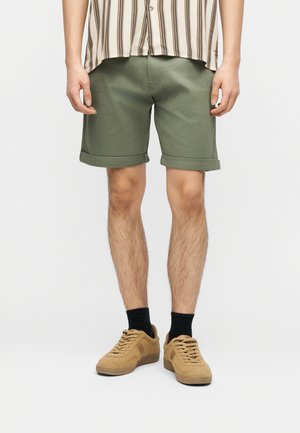 Mann trägt hochgekrempelte olivgrüne Shorts, schwarze Socken und beige Turnschuhe, steht vor einem schlichten weißen Hintergrund.