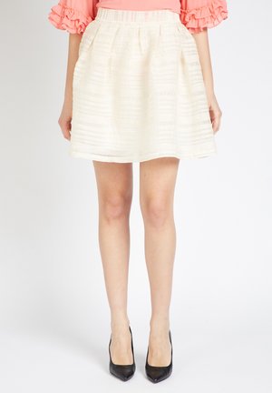 Mujer vestida con una falda acampanada blanca con textura, una blusa rosa con mangas acampanadas y tacones negros de punta, de pie contra un fondo blanco.