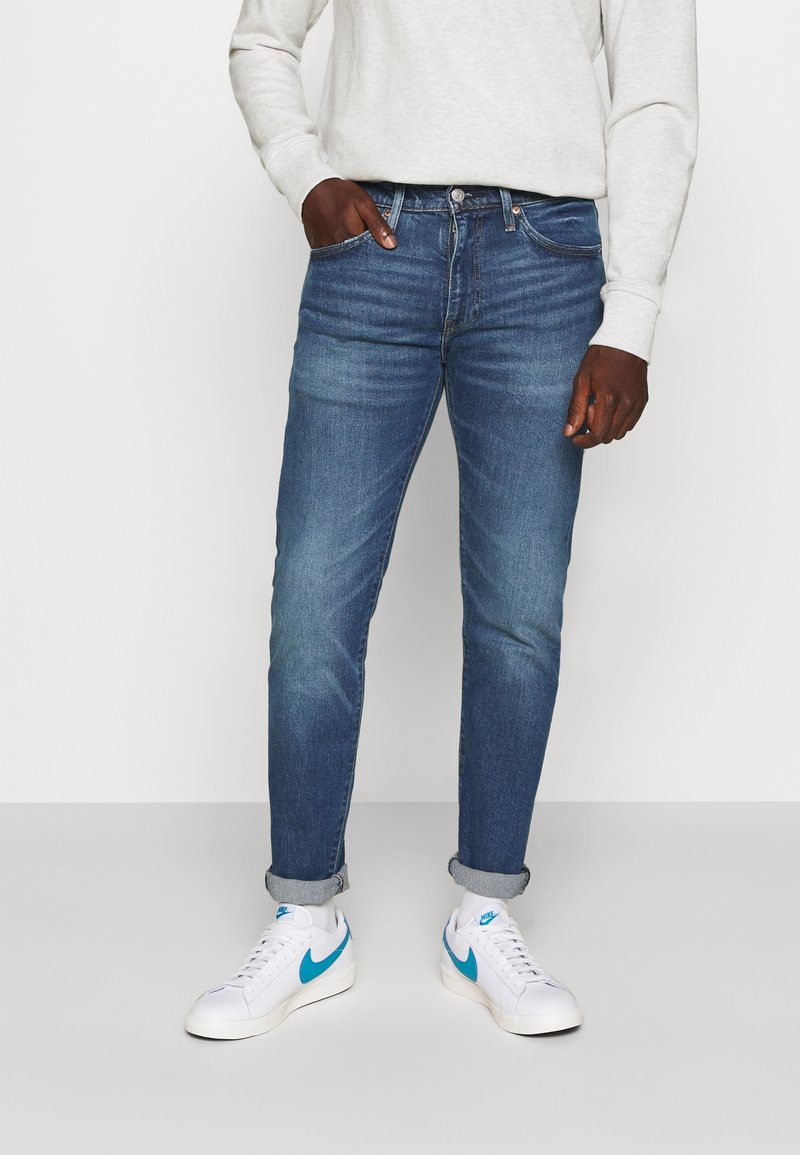 Homme portant un jean bleu retroussé, un sweat-shirt blanc et des baskets blanches avec un swoosh Nike bleu, debout avec une main dans la poche.