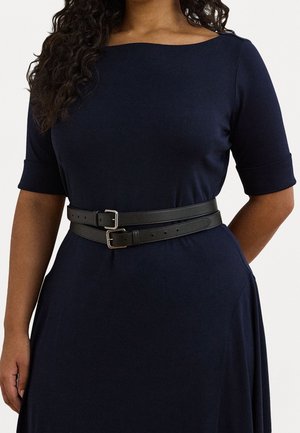 Kvinde iført en mørkeblå kjole med albuelange ærmer og et dobbelt sort læderbælte, der strammer taljen.