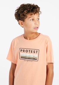 Perzikkleurig katoenen T-shirt met een rechthoekig zwart logo waarop "PROTEST" staat, met blauwe en groene strepen en een golfontwerp. Korte mouwen, ronde hals.