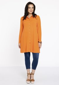 Orange skjorte med lange ærmer, buet kant og afslappet pasform, parret med blå skinny jeans og brune platform sandaler.