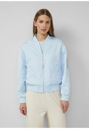 Frau trägt eine hellblaue Bomberjacke mit Reißverschluss, gerippten Bündchen und Kragen, kombiniert mit cremeweißen Hosen, stehend vor einem schlichten Hintergrund.