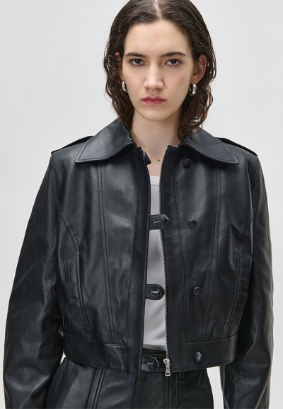 Mlada žena koja nosi crnu kožnu skraćenu jaknu s patentnim zatvaračem i kopčama preko bijele majice, gleda naprijed s neutralnim izrazom lica.
