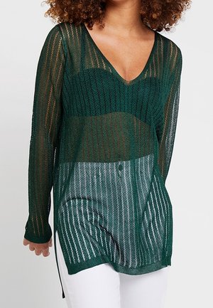 Femme portant un pull transparent à manches longues vert foncé avec col en V, par-dessus un bralette assorti, et un pantalon blanc, sur un fond uni.