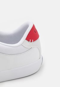 Baskets blanches avec une tige texturée, un accent rouge au talon et des coutures blanches. Forme arrondie avec une semelle en caoutchouc plate et un design minimaliste.