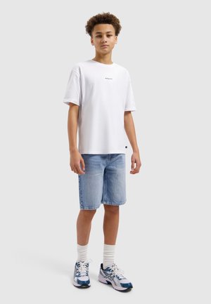 Tienerjongen staand, gekleed in een wit T-shirt, lichtblauwe spijkershorts, witte sokken en witte en blauwe sneakers, tegen een effen lichte achtergrond.