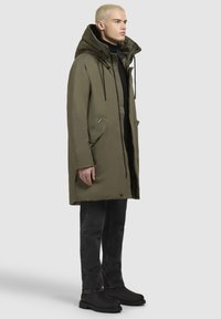 Olivenfarvet parka med hætte, der har lynlås foran, to sidelommer og sorte detaljer. Sorte jeans og kraftige sorte støvler fuldender looket.