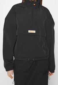 Czarna kurtka typu windbreaker z wysoką szyją, pół-zamkiem z przodu i elastycznym sznurkiem w dolnej części. Posiada biały prostokątny logo na klatce piersiowej.