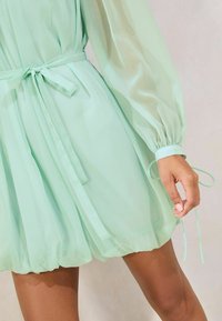 Rochie verde menta din material transparent, cu mâneci bufante, talie strânsă cu o fundiță și fustă evazată, stratificată.