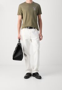 Olivgrön bomullst-shirt med logotyp, vita jeans i denim, svarta läderskor och en strukturerad svart väska med handtag. Enkel design, avslappnad stil.