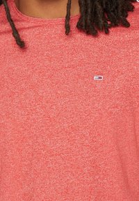 Primer plano de una persona que lleva una camisa de tela texturada roja con un pequeño logotipo rectangular y rastas cortas visibles.