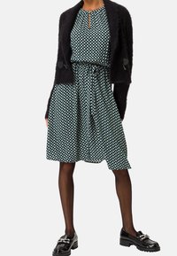 Tyrkysové šaty s bílým geometrickým vzorem, se staženým pasem, krátkými rukávy a černým huňatým cardiganem. Spárovány s lakovanými mokasíny.