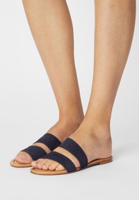 Piedi nudi che indossano sandali piatti a due fasce in suede blu navy con suole in pelle tan su uno sfondo bianco.