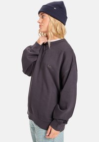 Donkergrijze oversized sweatshirt met een ronde halslijn en ribgebreide manchetten, voorzien van een klein logo op de borst en gecombineerd met lichte geruite broek.