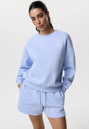 Kobieta ubrana w jasnoniebieską bluzę i pasujące spodenki, z rękami w kieszeniach, stojąca na jednolitym białym tle.