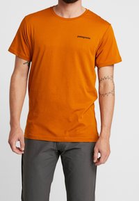 Mężczyzna w pomarańczowej koszulce Patagonia i szarych spodniach, pokazujący tatuaże na obu przedramionach, na jednolitym białym tle.