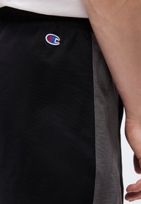 Πλάγια όψη κορμού ατόμου που φοράει μαύρο και γκρι σορτς με το λογότυπο Champion στο επάνω αριστερό μηρό.