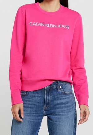 Personne portant un sweat-shirt Calvin Klein Jeans rose vif avec un jean bleu, debout devant un fond clair et uni.