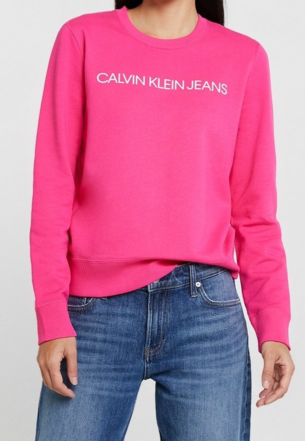 Personne portant un sweat-shirt Calvin Klein Jeans rose vif avec un jean bleu, debout devant un fond clair et uni.