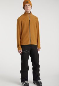 Braune Fleecejacke mit hohem Kragen, Reißverschluss vorne, getragen mit schwarzen wasserdichten Hosen mit Gore-Tex-Logo und Reißverschlusstaschen.