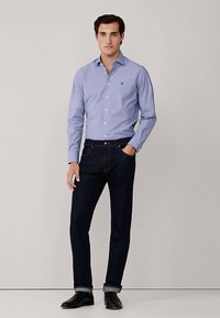 Ljusblå skjorta med knappar och en liten logotyp, kombinerad med mörkblå jeans. Jeansen har uppvikta fållar och matchas med svarta skor.