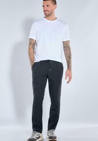 Man with tattooed arms wearing white t-shirt, black drawstring pants, and sneakers, standing against a plain light background.