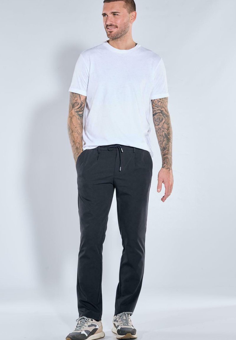 Man with tattooed arms wearing white t-shirt, black drawstring pants, and sneakers, standing against a plain light background.