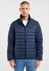 Jūras kara flotes puffer jaka ar rāvējslēdzēja aizdari un augstu apkakli, ar horizontāliem quiltētiem sekcijām un garām piedurknēm, kombinēta ar gaiši ziliem džinsiem.