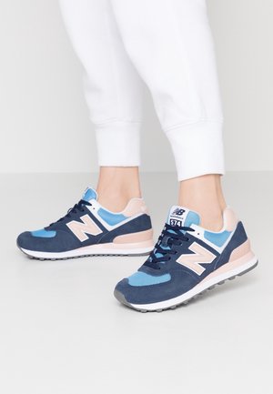 Person wearing navy blue, light blue, and beige New Balance 574 sneakers with white soles and white pants standing on a light grey floor.