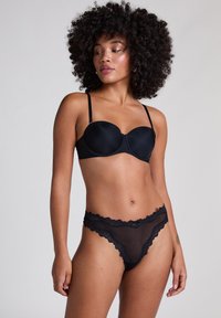 Zwarte beha van gladde stof met beugel, gecombineerd met doorzichtige zwarte kant slip met golvende randen. Model staat voor een neutrale achtergrond.