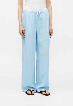 Nő világoskék, bőszárú, megkötős derekú nadrágban és fekete flip-flop papucsban, fehér háttér előtt áll.