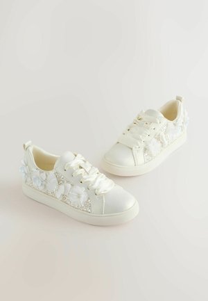 Białe trampki z satynowymi sznurówkami, błyszczącym bocznym panelem i kwiatowymi zdobieniami przy pięcie. Gładka cholewka i fakturowana podeszwa.