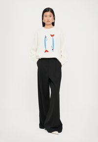 Witte gebreide trui met een blauw en rood geborduurd visontwerp, gecombineerd met hoge zwarte wijde broek en crèmekleurige schoenen.