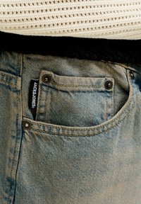 Vordertasche einer hellblauen Denim-Jeans mit sichtbarem schwarzem "Jack & Jones"-Etikett, getragen mit cremefarbenem Strickoberteil und schwarzem Kleidungsstück darunter.