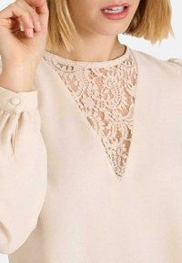 Blusa beige claro con un inserto de encaje en forma de V en el escote y mangas con fruncidos y detalles de botones. Textura de tela suave.
