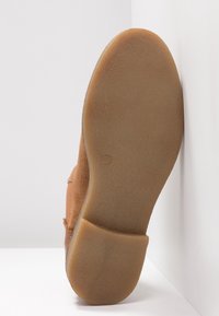 Bottine en daim marron montrant une semelle en caoutchouc texturée avec une légère courbe, inclinée contre un fond blanc, mettant en avant son design plat.