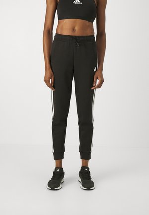 Osoba atletyczna w czarnym crop topie Adidas, czarnych spodniach dresowych z białymi paskami po bokach oraz czarnych butach sportowych Adidas, stojąca na białym tle.