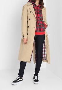 Trench coat beige con bottoni neri, indossato sopra una felpa a scacchi rossi e neri. Jeans scuri e sneakers bianche e nere completano l'outfit.