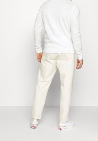 Weißer gestrickter Pullover mit langen Ärmeln, von hinten gezeigt, kombiniert mit hellbeigen Chinos und weißen Sneakers mit roten Akzenten.
