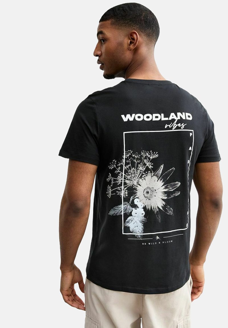 La camiseta de algodón negra presenta un estampado gráfico floral en blanco, que incluye el texto "WOODLAND vibes" y un diseño de borde rectangular.