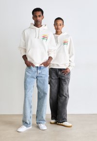Sudadera crema con bolsillo tipo canguro, que presenta bordados coloridos. Combinada con vaqueros de corte relajado en azul claro y gris oscuro, y zapatillas blancas.