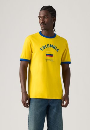 Uomo con i capelli intrecciati che indossa una maglietta gialla con rifiniture blu, scritta "Colombia", bandiera e jeans, in piedi davanti a uno sfondo semplice.