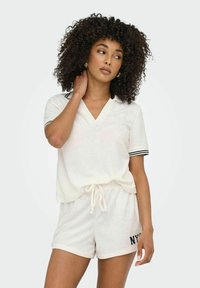 Cremefarbener Loungewear-Set bestehend aus einem V-Ausschnitt Oberteil mit kurzen Ärmeln und navyblauen Streifenakzenten sowie passenden Shorts mit dem Aufdruck "NYC". Weiche Textur.