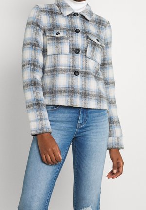 Persona con una chaqueta abotonada de cuadros azules, grises y blancos sobre un jersey de cuello alto blanco, combinada con vaqueros azules.