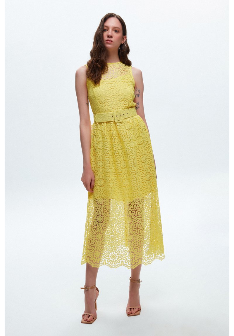 Gelbes Spitzenkleid mit tailliertem Oberteil, breitem Gürtel und floralen Mustern. Midi-Länge mit durchscheinendem, gewelltem Saum und schichtiger Textur.