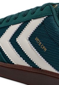 Grøn tekstilsneaker med hvidt chevron-design, syninger som detaljer og guldtekst. Har en brun gummisål og mønstret tekstur.