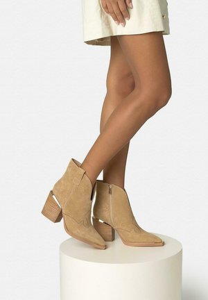 Femeie purtând cizme bej din piele întoarsă până la gleznă, cu tocuri suprapuse și detaliu cu fermoar, stând pe o platformă rotundă albă pe un fundal simplu.