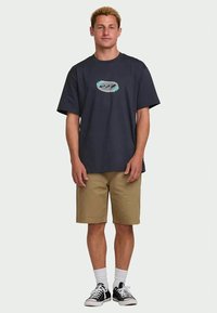 Young man standing wearing a navy graphic t-shirt, camel shorts, white socks, and black sneakers on a plain background.